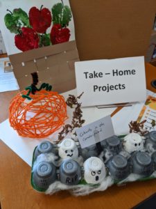 Weekly take-home projects for October on a desk with a sign reading "Take-Home Projects." Projects include apple stamping, a yarn pumpkin, a scavenger hunt and word game packet, and "ghosts in a graveyard" craft.